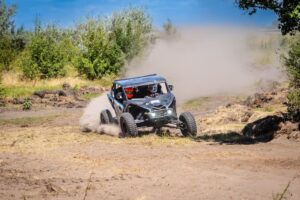 Off-road UTV or side-by-side vehicle racing through a dirt and grass track, kicking up dust.