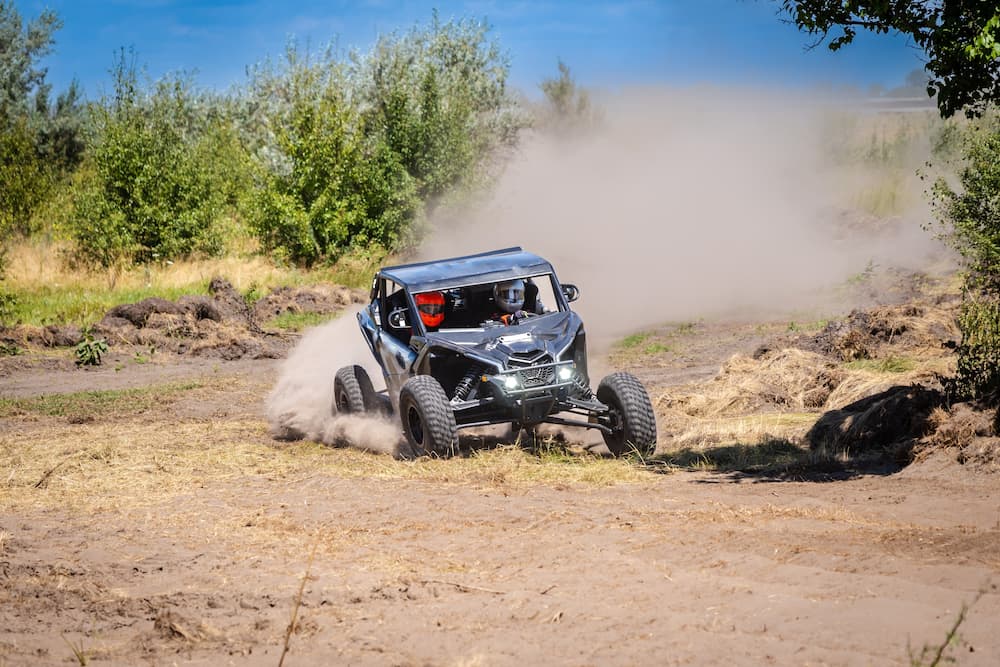 Off-road UTV or side-by-side vehicle racing through a dirt and grass track, kicking up dust.